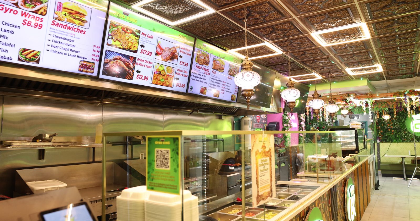 The Gyro King counter in Brooklyn, with a row of digital menu screens above the kitchen, an ornate pressed-metal ceiling and glowing hanging lanterns