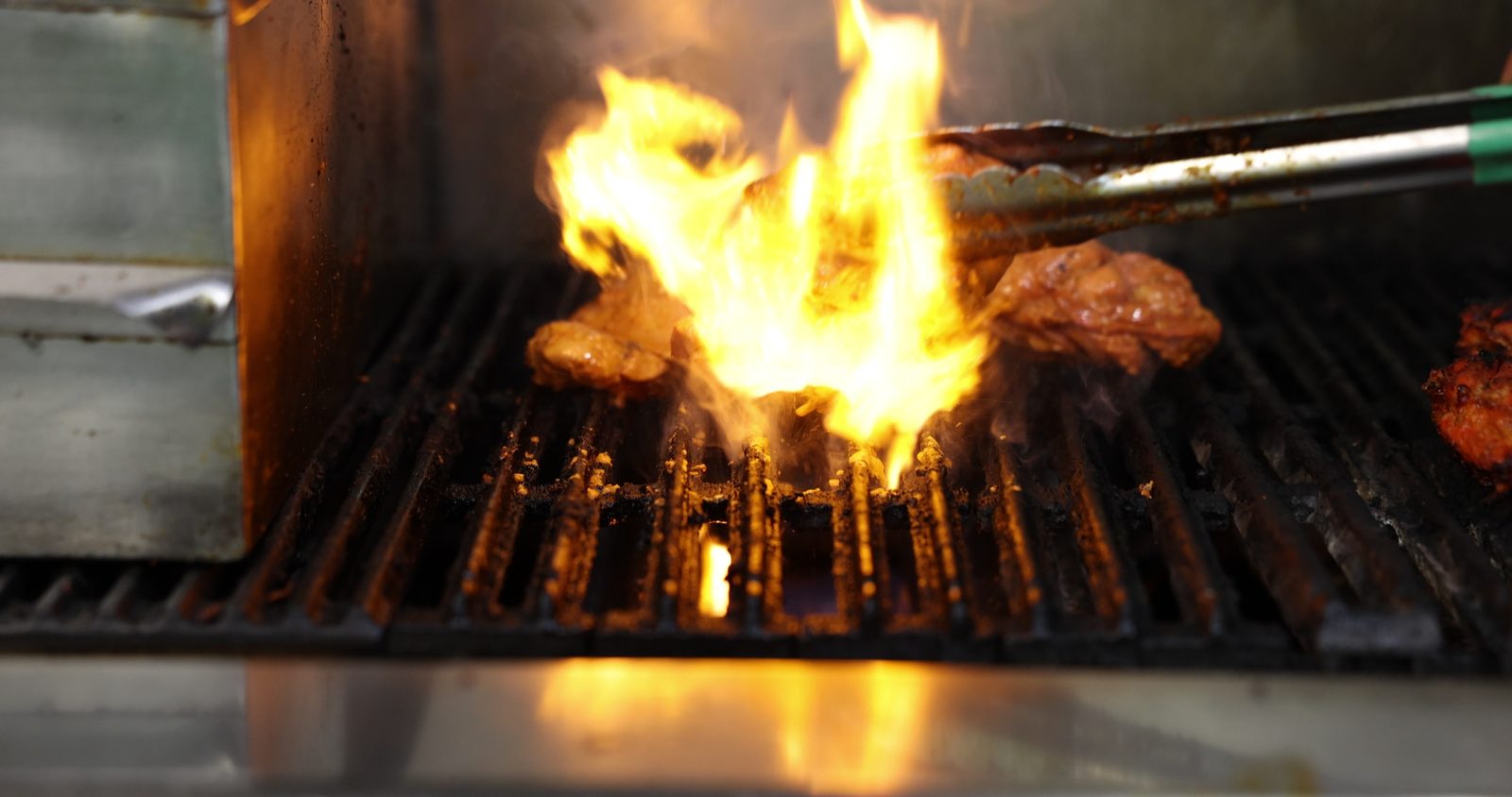 Marinated chicken on a restaurant grill with flames rising through the grates as tongs turn a piece, in the Gyro King kitchen