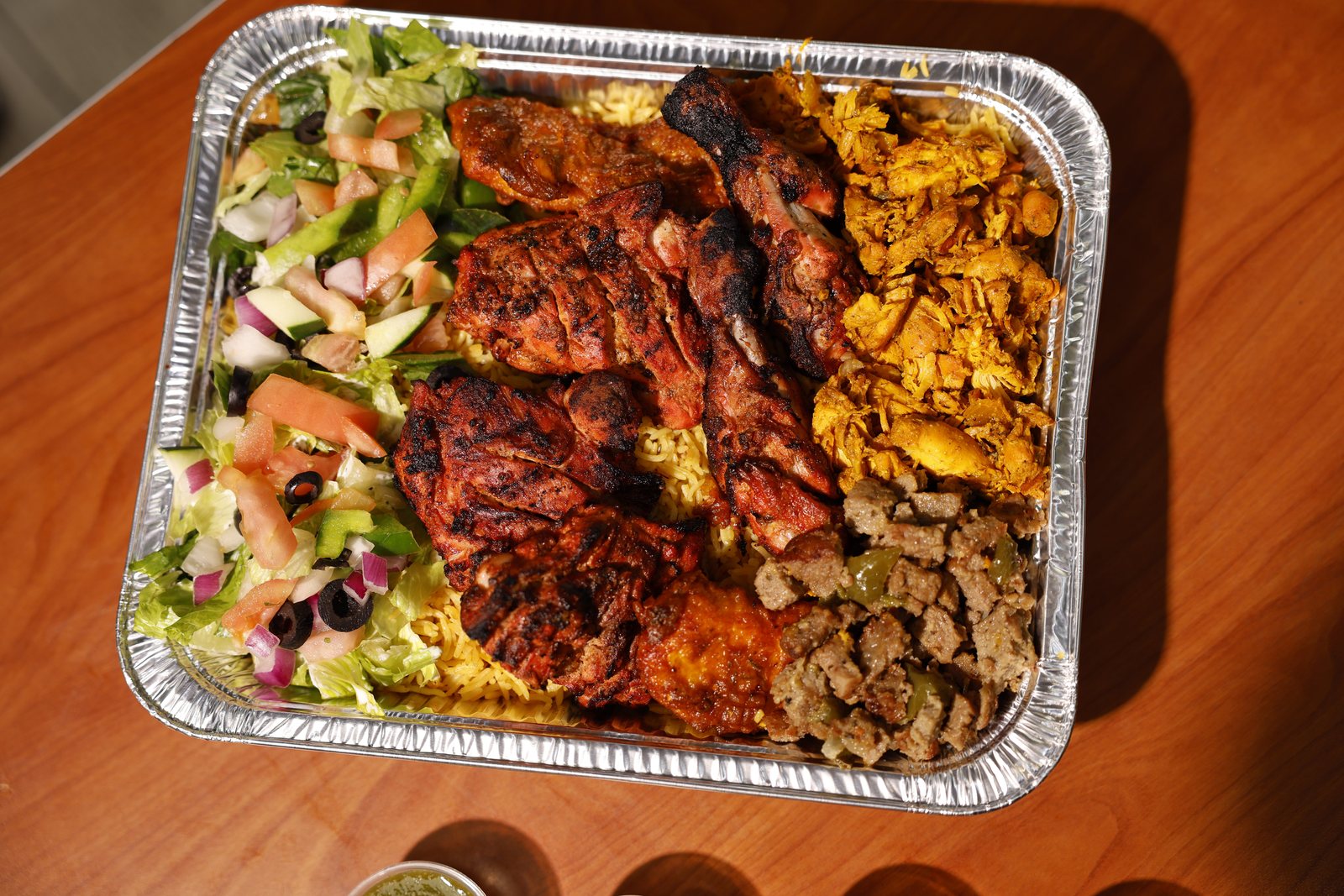 A Gyro King catering tray seen from above on a wooden table: grilled chicken, shredded chicken, seasoned beef cubes and yellow rice beside a fresh salad with olives and tomatoes