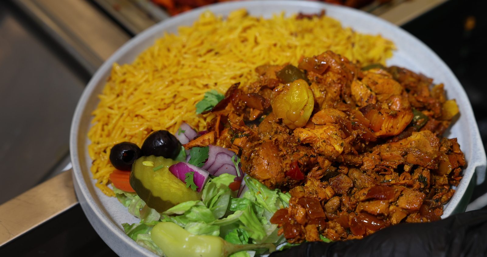 A gloved hand holding a fresh plate of chicken over yellow rice with salad, pickles and olives in the Gyro King kitchen